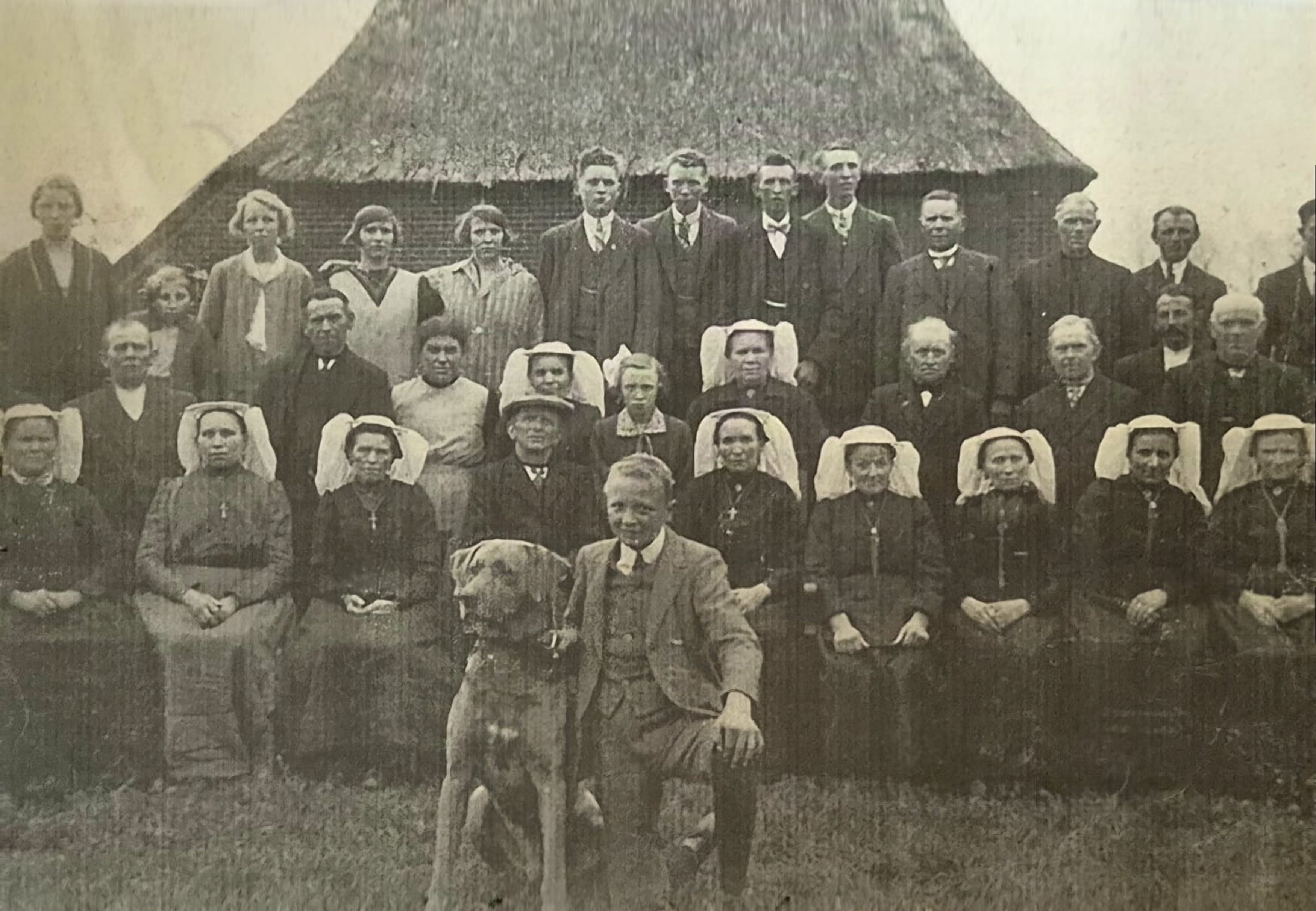 Oude groepsfoto bij een gebouw, met meerdere mannen en vrouwen en een hond
